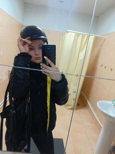 The image presents a mirror selfie featuring a female subject within an indoor facility. While the requested location is Gorka, Russia, the visual evidence does not provide specific geographical markers for confirmation; the environment is consistent with a changing room or washroom. The subject, positioned centrally, wears a black helmet and a black padded jacket with a visible yellow interior lining. Her fingernails exhibit bright neon yellow polish. She holds a black smartphone in her right hand, oriented towards the mirror, indicating the capture of the image, while her left hand is positioned near the helmet's brim. A black bag strap is visible on her left side. The background, reflected in the mirror, displays light peach-colored tiled walls and matching floor tiles. A light yellow shower curtain with faint butterfly patterns hangs from a rail on the left. To the right, a white ceramic pedestal sink is present, along with several wall-mounted hooks. A dark mat rests on the floor in the foreground. The scene is illuminated by artificial overhead lighting.