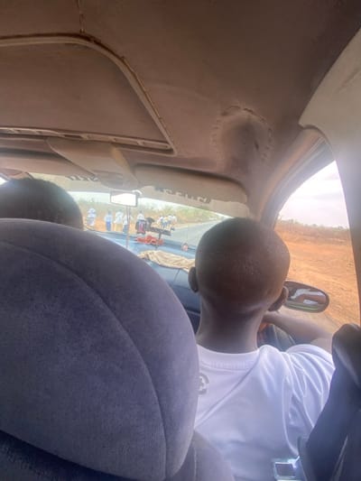 The image captures an interior view from a vehicle in Ninzo West, Nigeria, looking forward through the windshield. In the foreground, the back of a blue fabric passenger seat is visible on the left, and the back of a person's head, with short dark hair and wearing a white t-shirt, is positioned on the right, indicating a front-seat occupant. The vehicle's headliner shows visible stitching and signs of wear. A light-colored cloth is draped over a section of the dashboard. Outside the vehicle, a paved two-lane road extends into the distance. Approximately 10 to 15 individuals, predominantly dressed in white or light-colored clothing, are walking on the left shoulder of the road. The surrounding environment consists of dry, reddish-brown earth and sparse vegetation under a clear daytime sky. A vehicle is partially visible further ahead on the road.
