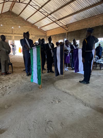 In a semi-enclosed structure with a corrugated metal roof and rough brick walls, a group of individuals in uniform are standing. Some are holding flags. One flag prominently displayed in the foreground is green, white, and green. Another flag nearby has black and white stripes, and further down, a flag with purple and white stripes is visible. The ground appears to be dirt. Several individuals are wearing dark uniforms with caps, some with white sashes. One person on the far left is wearing a beige outfit and glasses. The overall atmosphere suggests a formal gathering or ceremony, possibly a parade or a presentation event, taking place indoors. The city of Healtcare, Nigeria is not identifiable from the image itself, as there are no specific landmarks or contextual clues to indicate this location.