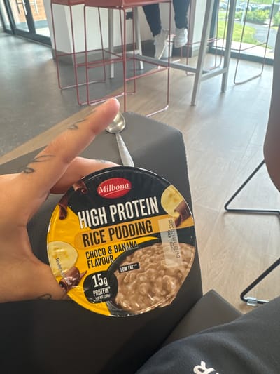 A hand with tattoos is holding a circular container of "Milbona High Protein Rice Pudding Choco & Banana Flavour." The container is yellow and black with text and images of banana and chocolate. A spoon is visible next to the container. The background shows a modern indoor environment with red and grey stools, a window, and a light-colored floor. There is no visual information in the image to determine a location within the city of Monaghan, Ireland.