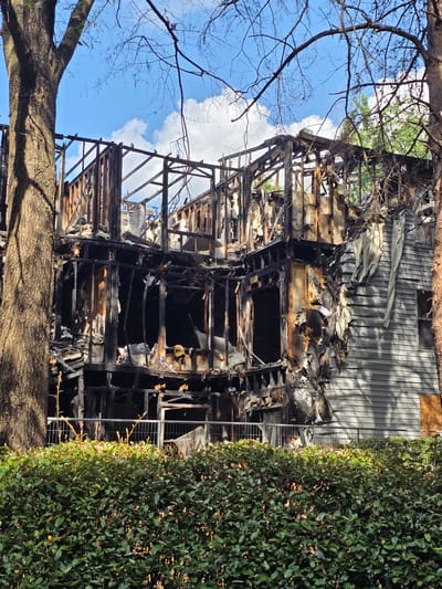 The image depicts a severely fire-damaged multi-story building, likely a residential structure, at 123 Oak Street in Roswell, United States of America. The front of the building has been almost entirely consumed by fire, with only the structural framework remaining. Charred wooden beams, blackened walls, and debris are visible throughout the exposed interior. Portions of the exterior siding on the right side of the building appear intact, although some areas are scorched and damaged. A light-colored vinyl fence runs horizontally across the foreground, separating the damaged structure from a dense, green hedge that occupies the bottom portion of the image. Bare tree branches extend from the tops of two large trees situated on the left and right sides of the frame, casting shadows on the scene. The sky above is partly cloudy, with a bright blue sky interspersed with white clouds. There are no people or animals visible in the image. The scene suggests a recent and destructive fire event.