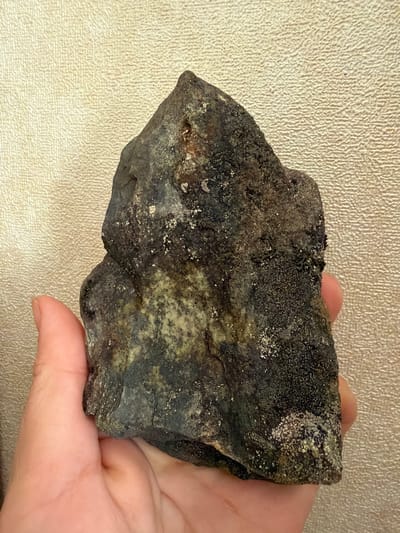 A hand holds a dark, irregularly shaped rock with a rough, textured surface. The rock exhibits patches of lighter green and numerous small, shiny, metallic flecks. It is held against a textured, pale gold background. The location is stated as Мяндиги, Russia, but this cannot be visually verified from the image alone.