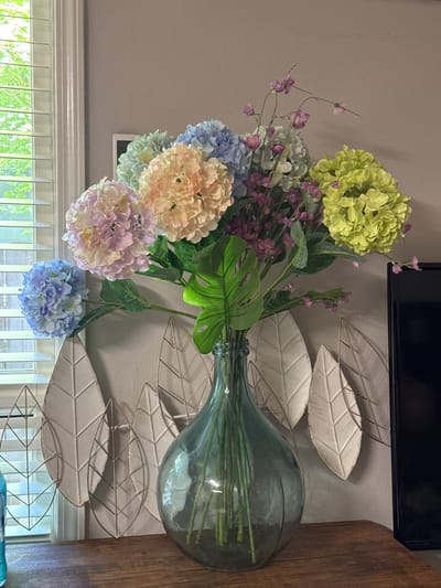 A close-up, eye-level shot captures a vibrant arrangement of artificial hydrangeas and filler flowers in a large, greenish-blue glass vase. The bouquet features a mix of soft pastel colors, including pale pink, peach, light blue, light green, and lavender hydrangeas, interspersed with delicate purple filler flowers and a large, textured green monstera leaf. The vase sits on a dark, wooden surface, likely a tabletop or shelf. Behind the vase, a neutral-toned wall serves as the backdrop, adorned with three cream-colored, leaf-shaped decorative wall hangings. To the left of the vase, a portion of a window with white horizontal blinds is visible, allowing natural light to filter through and illuminating the scene. To the right, a black rectangular object, possibly a television or monitor, is partially visible. The overall impression is one of domestic decor. The image does not contain any identifiable landmarks or features that would indicate a specific location within Vancouver, United States of America.