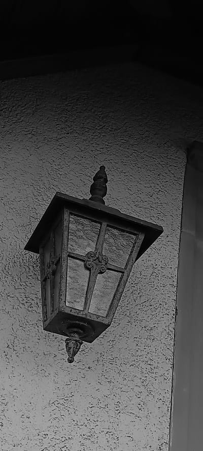 A black and white, low-angle, close-up shot shows an old, ornate lantern mounted on a textured, light-colored wall. The lantern is dark and appears to be made of metal, with decorative scrollwork in the center of each glass pane. The glass panes are frosted or textured, obscuring the view inside. A finial tops the lantern. To the right of the lantern, a vertical, darker column or beam is visible. The wall has a rough, stucco-like texture. The scene suggests an exterior setting, possibly a house or building entrance, in the city of Schwaikheim, Germany. No people are present.
