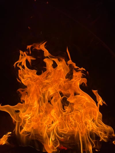 A vibrant, orange flame engulfs the right side of the frame against a black backdrop. The fire billows upwards and to the right, forming dynamic, swirling patterns. Wisps and tendrils of fire reach out, creating a sense of movement and heat. The contrast between the bright orange flames and the dark background highlights the intense glow and intricate details of the fire.