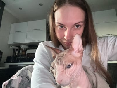 An indoor medium shot shows a young woman with long brown hair, wearing a white t-shirt, holding a light-pink, hairless cat in front of her chest. The cat, facing left, has blue eyes and wrinkled skin. The woman is smiling and looking directly at the camera. In the background, a white kitchen cabinet with silver handles is visible, along with kitchen appliances and shelves stocked with bottles. The scene appears to be in a domestic environment, possibly a kitchen, in Novaya Ivanovka, Russia.