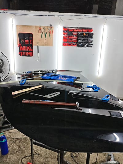 The image depicts a workshop environment, possibly for car repair or detailing, located in Krtsanisi, Georgia. The primary subject is a shiny black car hood, resting on a stand, with various tools laid out on its surface. These tools include what appear to be dent removal tools, a hammer with a wooden handle, and a set of specialized tools in a red and black case hanging on the wall. Behind the car hood, a white wall is visible, illuminated by bright LED strip lights. On the wall, a pegboard displays several pliers and screwdrivers. To the right of the pegboard, a wall-mounted organizer with multiple small red and black bins holds various small parts and tools. A large industrial fan is partially visible on the left side of the image. The floor is a rough concrete surface. A blue water bottle with a green handle is present on the floor near the car hood.