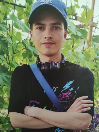 A young male is standing outdoors with his arms crossed in front of him. He is wearing a blue baseball cap, a black t-shirt with a graphic design, and headphones around his neck. A blue strap is visible across his chest. He is positioned in front of green foliage. The background suggests a natural or garden setting. The image is a portrait. The specific location in the city of Jablonec nad Nisou, Czechia is not discernible from the provided image.