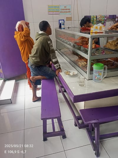 The image shows a scene in a food stall, located at latitude -6.655 and longitude 106.861, Kecamatan Ciawi, Indonesia. A young man, wearing a green shirt with white stars, sits on a purple wooden bench, leaning forward, his right hand resting on the table. A woman, wearing a bright orange shirt with white polka dots, stands beside him. She is holding her hand to her head, possibly in pain. A young man, wearing a black shirt and black pants, stands behind a counter, partially obscured. In front of him is a glass counter, filled with various food items. The counter is made of metal and glass. The image also shows two purple benches with a white table in between them. There is a glass of water and an empty plate on the table. A blue and white pitcher sits on the table to the right of the empty plate. There are various food items on display on the shelf behind the counter, including what appears to be fried chicken, chips, and other unidentified food. The background is a wall with faded purple paint. The scene appears to take place in a casual setting, possibly a street food stall. The date and time are captured as 2023/05/21 08:32.