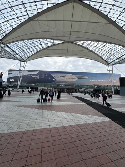 The image displays an outdoor concourse leading to Terminal 2 of Munich Airport, located in Oberding, Germany, under an overcast sky. A large, arching canopy, constructed from a metal grid supporting white fabric panels, covers the foreground and mid-ground. The ground is paved with rectangular tiles arranged in alternating patterns of light beige and reddish-brown, featuring a black drainage grate on the right side.
The background shows the multi-story glass and metal facade of Terminal 2. A prominent, large horizontal banner is affixed to the terminal building, displaying images of a dark blue Lufthansa Airbus A350-900 and a white Lufthansa Boeing 747. The banner also features the Lufthansa logo, "Star Alliance" emblem, and text including "A century since takeoff." and "100".
Approximately 20 to 30 individuals are present on the concourse, walking in various directions. Several pedestrians are observed pulling wheeled suitcases, indicating travel activity. On the far left, a small, dark brown, horned sculpture is positioned on a pedestal. Multiple white vans are visible parked near the terminal entrances in the distance.
