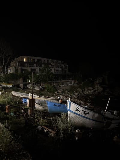 In Tyulenovo, Bulgaria, this image captures a night scene featuring a coastal landscape with a modern building and several boats. The building, likely a hotel or apartment complex, is multi-story with balconies and windows, one of which is lit. The foreground is occupied by a collection of boats resting on what appears to be a rocky or grassy embankment. Some boats are partially covered, while others are exposed. There are no people visible in the image. The environment is dark, with the only light sources being the illuminated window in the building and possibly some ambient light reflecting off the water or boats, creating a subdued atmosphere. The scene suggests a quiet, possibly off-season, coastal setting.