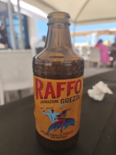 In San Salvo, Italy, a brown glass bottle with an orange label is positioned on a dark surface. The label features the word "RAFFO" in large red letters, with "LAVORAZIONE GREZZA" printed below it in smaller blue letters. An illustration of a blue figure spearing a blue creature is present on the label, along with text that reads "CEREALI DI PUGLIA NON RAFFINATI". The bottle is centrally located in the frame. The background is blurred, showing a bright, outdoor setting with glimpses of white structures, possibly tables and chairs, under a light-colored canopy. People are visible in the background, some appearing as blurred figures in motion. The overall scene suggests an outdoor dining or recreational area, likely a beachside establishment.