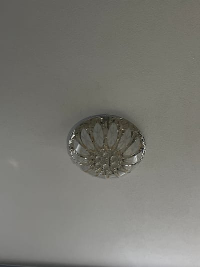 An upward-facing photographic image captures an interior ceiling in Bukovo, Bulgaria. The primary subject is a flush-mount or semi-flush-mount ceiling light fixture, positioned slightly off-center towards the lower-middle of the frame. This fixture is circular, featuring a polished silver-toned metallic base and a radial arrangement of translucent glass or acrylic elements. These elements are shaped like stylized petals or leaves, some frosted with etched linear patterns, surrounding a central cluster of multiple small, clear spherical crystal or glass adornments. The ceiling itself is a flat, light grey or off-white surface with a discernible textured finish. No individuals, overt actions, or interactive elements are present in the scene.