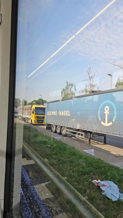The image captures a view through a window, likely from a vehicle or building, showing a street scene in Neu-Ulm, Germany. In the foreground, a grassy embankment runs alongside what appears to be a railway track. Further back, a yellow semi-truck is visible, followed by another semi-truck with a blue trailer. The blue trailer has the words "KUEHNE+NAGEL" emblazoned on its side, along with a large anchor logo. The sky is a bright blue with scattered clouds, and sunlight illuminates the scene, casting soft shadows. Trees and a street lamp are also present in the background, suggesting an urban or suburban environment. The overall impression is of a typical roadside scene during daytime.