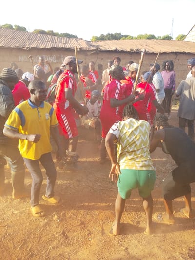 The image depicts an outdoor gathering of approximately twenty individuals in Vodni, Nigeria, under daylight conditions. The scene is set on unpaved reddish-brown dirt ground, with visible dust suspended in the air. In the background, a single-story structure with a corrugated metal roof and a light-colored wall features a partially visible inscription "NO FIGHTING." A group of at least eight individuals centrally located wear matching red short-sleeved shirts with white shoulder stripes and red bottoms with white side stripes. Several in this group hold slender wooden staffs and appear to be moving rhythmically, generating dust. To the left foreground, a man in a yellow and blue polo shirt, dark pants, and yellow slides gestures with his left arm while stepping. To the right foreground, another individual, viewed from behind, wears a yellow and white patterned shirt and green shorts, crouched with hands slightly raised. Other individuals in varied attire are present around the perimeter, observing or participating. A person seated on the ground near a small drum is visible in the middle ground.