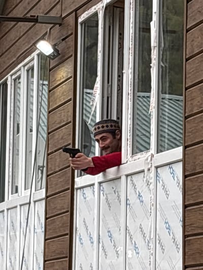 The image shows a man holding a gun while looking through a window. The man is wearing a red shirt and a patterned cap. The window is made up of multiple panes, some of which have a protective film with "PANEL TECH" printed on them. The location is possibly Kveda Chkhutuneti, Georgia, but this is inferred from metadata. The environment appears to be an interior with a roof structure. A light fixture is visible in the lower-left corner.