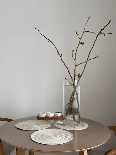 The scene depicts an interior setting in the city of Башкултаево, Russia, characterized by a minimalistic aesthetic. A round, light-colored wooden table occupies the mid-ground, with partial views of two matching wooden chairs flanking it. A plain, light grey wall serves as the background.
Centrally positioned on the table is a tall, clear cylindrical glass vase filled with water. Inside, several bare tree branches with emerging green buds are arranged, their lower ends submerged and anchored by a dark, irregular object at the bottom of the vase. Condensation is visible on the outer surface of the vase.
To the left of the vase, three circular woven placemats, light beige in color, are laid on the table. One is directly beneath the vase, and two are positioned in the foreground. Behind these placemats and to the left of the vase, a small rectangular wooden tray holds three small, handleless ceramic vessels: two are white, and one is dark grey. No individuals or animals are present, and no actions or interactions are depicted within the frame.