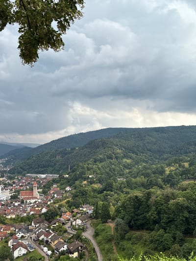 This image captures a scenic view of Gernsbach, Germany, from a high vantage point. The scene is dominated by a dense, green forest covering a steep hillside that slopes down towards a town nestled in the valley. The town features numerous houses with red-tiled roofs, clustered along winding roads. A prominent church with a tall steeple is visible within the town. The weather appears overcast, with dramatic, heavy grey clouds filling the upper portion of the frame, contrasting with the lush greenery below. A large tree with green foliage occupies the lower left corner of the image. The overall impression is one of a natural landscape meeting a quaint European town under a moody sky. The time of day is not explicitly clear but suggests daytime. No text is visible in the image.
