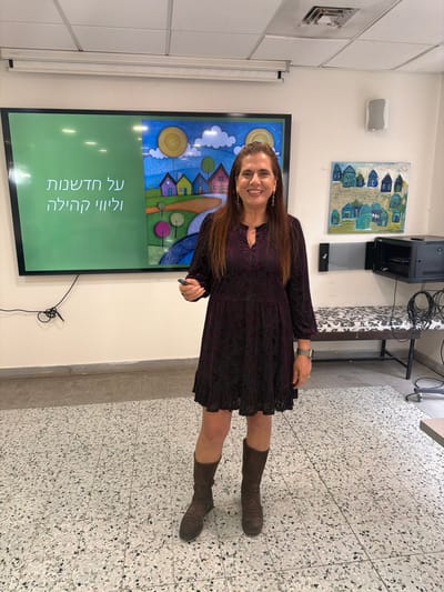 A woman stands in front of a large screen with text in Hebrew that translates to "About news and community". The screen displays colorful graphics of houses under a sun and clouds. The woman wears a dark, long-sleeved dress and brown boots. Behind her, on the wall, are a speaker, another picture, and a small table. The floor has a speckled pattern. The room appears to be a meeting or presentation space.