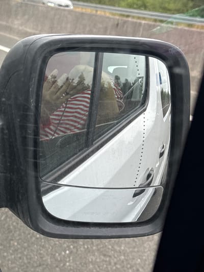 The image shows a reflection in a car's side mirror of the interior of a white van. Inside the van, a person wearing a red and white striped shirt is visible. Their hand, covered in white material, is raised near their face. An American flag is also visible inside the van, hanging from the window. The van is driving on a road, indicated by the asphalt and the highway barrier visible in the background. The image appears to be taken in Dunbell, Ireland.