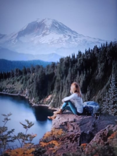 The image depicts a solitary figure, a person with blonde hair, sitting on a rocky ledge overlooking a body of water. The individual is wearing a light-colored top and teal-colored pants, with hiking boots. Next to them is a blue backpack. The background features a dense forest of evergreen trees, with a snow-capped mountain range in the distance under a hazy sky. The foreground shows some autumn foliage and foliage on the rocky ledge. The scene is a natural landscape, likely a mountainous region. There is no indication that this scene is located in the city of Balakliya, Ukraine.