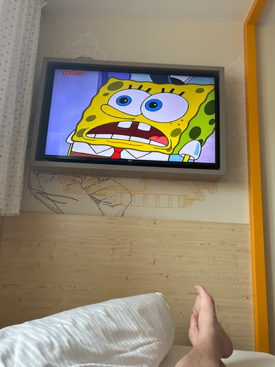 A brightly lit interior room features a mounted flat-screen television displaying an image of Spongebob Squarepants. The TV is framed in gray and positioned against a wall with patterned wallpaper depicting sketched architectural or industrial designs. Adjacent to the patterned wallpaper is a section of light wood paneling. In the right portion of the image, a person's foot is visible, extending towards a white pillow and bed. The room also has a bright yellow decorative molding at the top of the wall. The exact location within Ingolstadt, Germany cannot be determined from the given image.