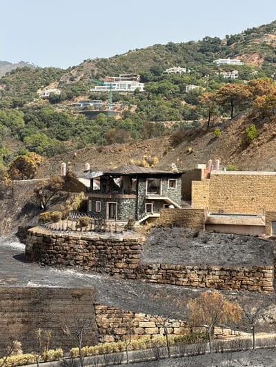 The image depicts the aftermath of a wildfire in a hillside residential area near Montemayor, Spain. A two-story house with a stone facade and a damaged roof is prominently featured, showing signs of extensive fire damage, including blackened walls and a collapsed roof section. The surrounding landscape is scorched black, with burnt vegetation and ash covering the ground and terraced stone walls.
In the background, several modern white houses are visible on the slopes, some appearing intact and others potentially affected. A green construction crane is also present on a distant hilltop, suggesting ongoing development in the area. The terrain is characterized by steep, dry hillsides dotted with green trees and shrubs, contrasted by the stark blackness of the burnt areas. The sky is clear and bright, indicating a daytime scene.