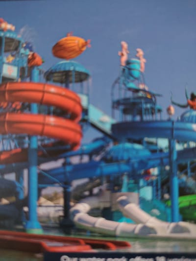 The image depicts a water park with several brightly colored slides and structures. In the foreground, large red spiral slides are prominently visible, supported by blue pillars. The background features a complex network of blue slides and platforms, with a distinctive orange blimp-like object floating in the sky above the water park. There are also two pinkish figures on top of a blue structure in the upper right portion of the image, possibly decorative elements or part of a ride. The sky is a clear blue. The image is slightly out of focus, giving it a soft appearance. The text at the bottom of the image indicates it is related to a water park, possibly a promotional image. There is no visible indication of the city of Bahshitpa, Uzbekistan in the image itself.