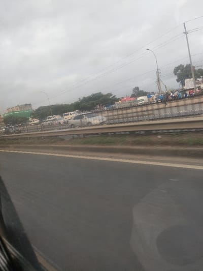 The image depicts an elevated view of a multi-lane asphalt highway, likely in Kiuu ward, Kenya, under an overcast sky. In the immediate foreground, a dark, angled object, consistent with a vehicle's interior component, partially obscures the lower left corner, indicating the photograph was taken from a moving vehicle. The primary roadway in the midground features a grey asphalt surface with a faint yellow line at its right edge. This road is separated by a grassy median and a concrete barrier (jersey barrier style) topped with a metal guardrail from an elevated service road or adjacent thoroughfare.
On this elevated section, multiple vehicles are visible, including a blurred silver sedan, white vans, and other cars, suggesting active traffic. Beyond the traffic, a crowd of people is present along the roadside, accompanied by various makeshift structures or stalls with colorful tarpaulins (red, blue), indicative of informal vending or a market area. A large utility pole with overhead power lines and a street light fixture is positioned above this elevated section. In the distant background to the left, a multi-story building, possibly under construction and partially covered in green netting, is discernible amidst trees. The environment includes numerous green trees bordering the roadways.