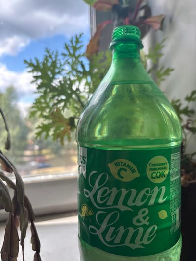 A green plastic bottle of "Lemon & Lime" soda, containing a clear liquid, is positioned on a windowsill. The bottle has a label with the text "VITAMIN C" and "Содержит лимонный СОК". In the background, through the window, a cloudy blue sky and green foliage are visible. The location is identified as Tutaev, Russia, based on the inscription on the bottle which reads "г. Самара", which is incorrect. The bottle actually has a manufacturing address in Tutaev, Russia, indicating the place of origin of the beverage. The context of Tutaev, Russia, is inferred from the label's printing origin which appears to be from that city.