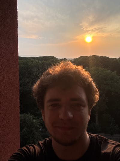 A young man with brown, curly hair, illuminated by the setting sun, takes a selfie outdoors. He is wearing a dark t-shirt. To his left, a textured red wall is visible. In the background, a dense forest stretches towards the horizon, with a bright, setting sun peeking through the hazy sky above it. The scene appears to be in or around Kolomyia, Ukraine, as indicated by the provided context.