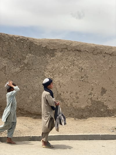 The image depicts two young boys walking along a dusty path next to a tall, textured mud wall under a partially cloudy sky. The scene suggests a rural setting in daylight, consistent with the provided context of Panjwai, Afghanistan.

The boy in the foreground, positioned right of the center, walks towards the right side of the frame. He wears a light tan or beige shalwar kameez, a white and black turban-like head covering, and a dark blue scarf around his neck. He has light-colored sandals on his feet and carries a bundle of fabric, possibly clothes, in his left hand. His gaze is directed forward.

Behind him, closer to the left edge, a second boy also walks towards the right. He wears a light blue or grey shalwar kameez and is barefoot. His right hand is raised to touch a white skullcap or small turban on his head, a gesture that could be adjusting it or shielding his eyes from the sun.

Both boys cast clear shadows on the ground, indicating a bright, sunny day. The path they walk on appears to be a dirt shoulder adjacent to a narrow strip of paved road or concrete. The dominant background feature is a high, rugged wall made of earth or adobe, showing rough textures and some erosion. Above the wall, a pale blue sky with wispy white clouds fills the upper portion of the frame. There is no visible text in the image. The boys' expressions are neutral, reflecting a sense of routine activity.