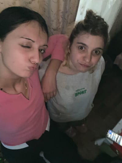 Two females are captured in a selfie-style photograph indoors, set in the city of Lanchkhuti, Georgia. The image is taken from an elevated perspective. The female on the left, with dark hair, is wearing a pink V-neck top over a white undershirt and dark trousers. She winks her left eye and puckers her lips, while her right arm extends around the shoulder of the second female. A thin, gold-toned necklace is visible on her neck. The female on the right has light brown hair arranged in a top knot and is dressed in a light grey long-sleeve shirt with "HOLLOGYN SPORT" printed in green on the front; dark moisture stains are present on the fabric. She maintains direct eye contact with the camera, exhibiting a subtle smile. The background features a light-colored, patterned curtain on the right side and indistinct, dimly lit interior walls. A portion of a smartphone screen, displaying an active interface, is visible in the lower right corner of the frame.