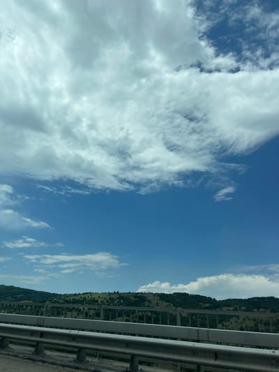 This image captures a wide-angle view of a natural landscape and human-made infrastructure in Gorno Varshilo, Bulgaria. The upper two-thirds of the frame are dominated by a dynamic sky, featuring a blend of dense, white-to-grey stratocumulus clouds in the upper left transitioning to patches of clear, deep blue sky and scattered lighter cumulus and cirrus clouds towards the right and middle. The horizon line is visible in the lower third, revealing rolling hills covered with varied vegetation. These hills are predominantly green, indicating lush growth, with a mix of grassy areas and numerous scattered trees or small clusters of woodland. In the immediate foreground, a substantial metallic road barrier or bridge railing spans the width of the frame. This barrier consists of multiple horizontal silver-colored rails secured by visible bolts and supported by vertical posts, anchored into a concrete structure. The concrete base shows signs of wear and minor debris. No people, animals, or vehicles are present in the scene.