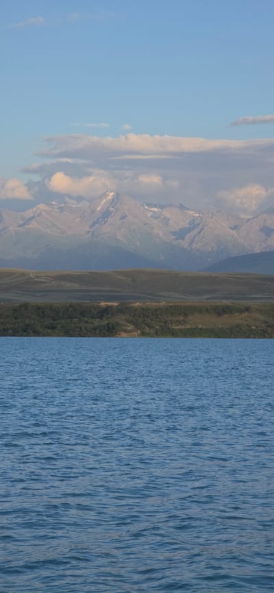 The image captures a serene natural landscape at Pristan'-Przheval'sk, Kyrgyzstan. In the foreground, a vast expanse of calm blue water stretches across the frame, its surface rippled by a gentle breeze. The water occupies the lower half of the image, creating a sense of depth and tranquility.
Above the water, a verdant green hillside rises, covered in dense vegetation that appears as a dark carpet. The hillside is textured with rolling contours and subtle variations in color, suggesting undulation and depth.
Further in the background, majestic mountains dominate the horizon. These peaks are rugged and imposing, with some of their summits capped with snow, gleaming white against the sky. The mountains exhibit a rugged, layered appearance, with sharp ridges and deep ravines, indicating a grand geological formation. The sky above is a pale blue, with scattered wispy clouds that add a touch of softness to the scene. The light suggests it is either early morning or late afternoon, casting a gentle glow over the landscape.
There are no people or man-made structures visible in the image, emphasizing the raw, untamed beauty of the natural environment. The overall impression is one of peace and grandeur, a moment frozen in time showcasing the natural splendor of the region.