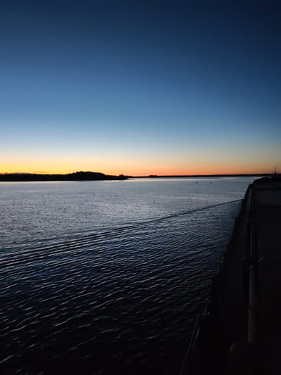 An outdoor, vertical, wide-angle shot taken at dusk from a vessel looking out over a large body of water. The sky transitions from a deep blue at the top to a pale orange-yellow glow along the horizon. The water is dark and rippled, with a wake trailing from the right side of the frame, indicating movement. In the distance, a dark, tree-lined landmass is visible on the left horizon. On the right side of the frame, the dark, textured edge of the vessel is present, with some vertical poles. The scene suggests a serene, twilight moment over water.