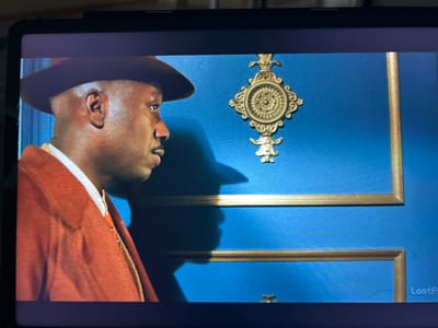 A man wearing a hat and an orange coat stands in front of a blue wall with gold trim. A decorative gold ornament is attached to the wall. A shadow of the man's head and hat is cast on the wall. There is text that says "LostFi" in the bottom right corner. The location is not specified in the image.