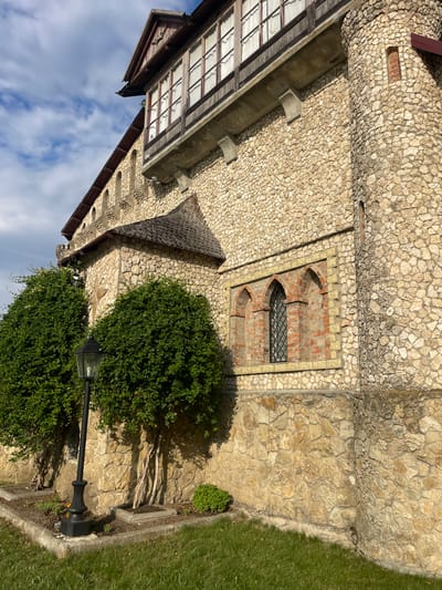 The image displays a detailed view of a rustic stone building, resembling a castle or old manor, in Colibași, Moldova. The building features walls constructed from irregular, light-colored stones, with sections of red brickwork visible around arched windows. One prominent window on the right has three Gothic-style arches and is fitted with a black metal grate. Above this, a balcony with wooden framing and numerous glass panes stretches along the upper level of the building. A dark, pitched roof, likely made of shingles, covers a portion of the structure. In the foreground, well-maintained green bushes flank a black lamppost with a clear glass lantern. The ground is covered in green grass, with small planted areas around the bushes and lamppost. The sky above is a mix of blue and white clouds, suggesting it is daytime. The architectural style and the surrounding vegetation evoke a historical and somewhat secluded atmosphere.