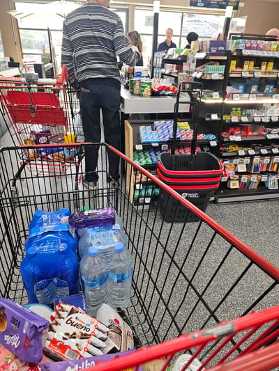 This image provides a first-person perspective from within a black wire shopping cart with red handles, situated inside a convenience store or small supermarket in Valence, France. The store features a speckled dark grey floor and is brightly illuminated by overhead lighting and additional task lighting on shelves.
The foreground of the shopping cart contains two multi-packs of 1.5-liter bottled water (one with blue plastic wrapping, labeled "Plafena"; another with clear plastic wrapping, labeled "Source Alpine"), several chocolate products including Milka "Choc & Choc" and Kinder Bueno multi-packs (6x43g), a white bag from "La Boulangere," and a small purple bottle resting on the water packs.
Ahead, at the checkout counter, a man is seen from the back, wearing a horizontally striped long-sleeve shirt and dark blue jeans with a wallet partially visible in his right back pocket. He is standing at a light-colored counter, presumably making a purchase. A female cashier with light brown hair is partially visible behind the counter, attending to him. To the man's left, a red wire shopping cart, equipped with a small fan on its handle, contains a yellow bottle and other items.
In the background, behind the checkout, a man with short dark hair in a dark shirt stands near large glass windows that reveal an outdoor area with a white umbrella. Another person's head is partially visible on the far right.
Above the checkout counter, two vertical lighted pillars are numbered "7" and "8," with a digital display between them showing "13" and "20H39." The counter displays various small items and includes canned goods, such as one labeled "LABELL." To the right of the checkout, black shelving units extend along the wall, stocked with numerous snacks, candies (e.g., FULFIL, Skittles, Mentos, Milka, Kinder), and drinks. Below these shelves, a stack of black "SHOP & ROLL" branded shopping baskets with red handles is visible.