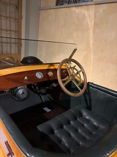 An interior view of a meticulously preserved vintage automobile is presented, featuring a classic design with rich materials. The image, originally rotated 90 degrees counter-clockwise, primarily focuses on the driver's area and a portion of the passenger seating.
The prominent feature is a wooden steering wheel with intricate brass spokes and central controls, indicative of early automotive engineering. Below the steering wheel, a polished wooden dashboard or panel extends, crafted from a combination of lighter wood trim and a darker, possibly mahogany, section. Embedded in this panel are two circular metallic emblems or gauges; one appears to be a crest, while the other features a stylized winged figure, possibly an early brand logo. A small metallic knob or switch is also visible on the panel.
Adjacent to the driver's area is a plush, dark grey or black tufted seat, likely upholstered in leather or high-quality faux leather, exhibiting deep buttoning and fine craftsmanship. A dark, textured floor mat covers the footwell area.
The background suggests an indoor setting, possibly a museum, showroom, or private collection in Wassenaar, Netherlands, given the clean condition of the vehicle and the surrounding architecture. A light-colored, mottled wall or ceiling is visible above, along with a portion of a lighter grey wall and a wooden structure with multiple glass panes in the lower left, hinting at a display cabinet or architectural feature within the building. A thin, dark rod extends upwards from the car's interior into the background. There is no clearly legible text visible on any of the car's components or the surrounding environment.