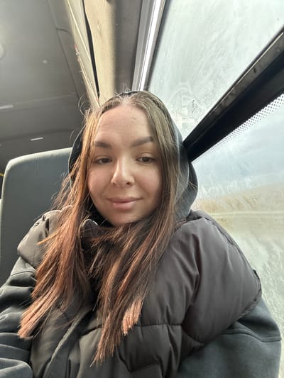 A woman with long brown hair is shown in a selfie inside a vehicle, likely a bus or van, based on the overhead window and interior details. She is wearing a dark, puffy jacket with a hood. The window above appears to be frosted or dirty. The seat next to her is grey. The location in Bol'shaya Sosnova, Russia is inferred from the prompt.