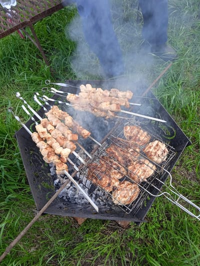 An outdoor barbecue is taking place on a grassy area, likely in Тайсуганово, Russia. A black metal brazier contains glowing coals and ash, upon which various meats are being grilled. Several skewers of pork chunks are positioned over the coals, and a metal grill basket holds pieces of grilled chicken. Smoke rises from the barbecue, indicating the cooking process. In the background, the lower legs of a person wearing blue pants are visible, suggesting their presence at the barbecue. The surrounding environment is lush green grass, and part of a rustic metal grill stand is also visible. The overall scene depicts a casual outdoor meal preparation.