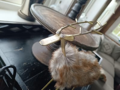 A close-up, eye-level shot focuses on a small, decorative reindeer figurine, its body covered in soft, fluffy fur that transitions from light grey at the top to reddish-brown at the bottom. The reindeer has delicate, brass-colored antlers that are thin and branch out like twigs. The figurine is positioned in the foreground, obscuring much of the background.

Behind the reindeer, a dark, glossy surface, likely a table or shelf, is visible. On this surface rests a wooden object with a raised rim, appearing to be a platter or a shallow bowl. The wood has a visible grain and a warm, brown tone. Further in the background, out of focus, are hints of a window with natural light streaming in, suggesting daytime. Beyond the window, blurred greenery can be seen, implying an outdoor setting, possibly with trees.

To the left of the wooden platter, a tall, metallic object with a curved base and a dark, cylindrical top is partially visible. This object has a sleek, modern design. The overall scene has a warm, indoor feel, with soft lighting creating a cozy atmosphere. The focus is squarely on the detailed craftsmanship of the reindeer figurine, highlighting its textures and metallic accents. There is no visible text in the image.