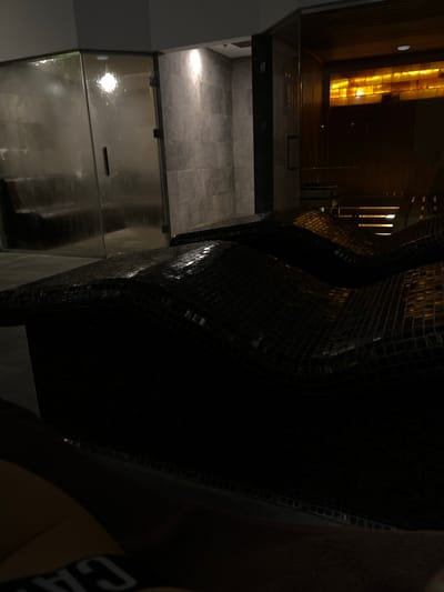 The image shows the interior of a sauna. There are curved structures covered in small, dark tiles. A glass door leads into a tiled room. Light emanates from a panel on the ceiling, suggesting a warm or relaxing ambiance. The photo appears to be taken in an indoor setting. Пампорово, Bulgaria is mentioned as a possible location.
