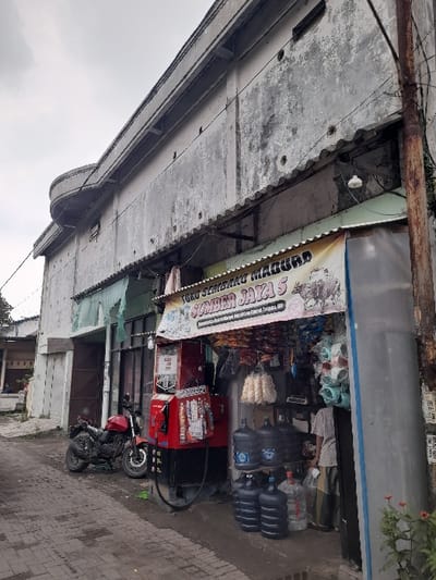 The image captures an outdoor scene in Kecamatan Gempol, Indonesia, focusing on a small storefront and the immediate surroundings under an overcast sky. The photograph is oriented vertically, requiring a 90-degree clockwise rotation for proper viewing.
The main subject is a small shop, likely a convenience store or 'sembako' (staple goods) shop, with an awning extending over its entrance. The awning is yellow and white, with prominent red and black lettering that reads "TOKO SEMBAKO MADURA" and below it, "SAMPAI JAYA S." Inside the shop, various goods are visible, including multiple large blue and black water dispenser bottles (gallons), bags of snacks or crackers, and other packaged items on shelves. A red machine, possibly a beverage cooler or a fuel dispenser for small quantities, is also visible near the front.
To the left of the shop, a concrete building with a somewhat weathered facade extends into the frame. Overhead, thin wires, possibly electrical or communication lines, run along the exterior of the building. The ground in front of the shop and to the right is paved with interlocking bricks or cobblestones, suggesting a sidewalk or a narrow street. A red and black motorcycle is parked directly in front of the shop's entrance, facing right. Another red machine, possibly a small fuel pump or a vending machine, is positioned near the motorcycle.
The overall scene depicts a typical modest commercial area, likely in a residential neighborhood, with a small business catering to local needs. No people are visible in the frame, and the time of day appears to be daytime, given the ambient light, though the overcast sky prevents direct sunlight.