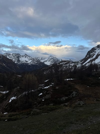 The image presents a vast, rugged mountainous landscape in Poschiavo, Switzerland, under a dramatic sky. Snow-capped peaks dominate the background and mid-ground, their jagged forms partially veiled by dynamic cloud formations. The sky features a mix of heavy, dark grey clouds and brighter, illuminated patches of white and light blue. A small, distinct moon is visible in an open, lighter section of the sky, suggesting either dawn or dusk.

The mid-ground reveals slopes dotted with dark, bare trees, indicative of late winter or early spring, with remnants of snow cover on the ground. The foreground consists of uneven, dark terrain comprising earth, rocks, and sparse, dry vegetation or grass, also with scattered patches of snow. Strong shadows are cast across the foreground and certain mountain slopes, indicating a low-angle light source, likely a sun hidden behind the clouds or mountains. There is no human activity, objects, or text visible in the scene. The overall impression is one of a grand, tranquil, and cold natural environment.