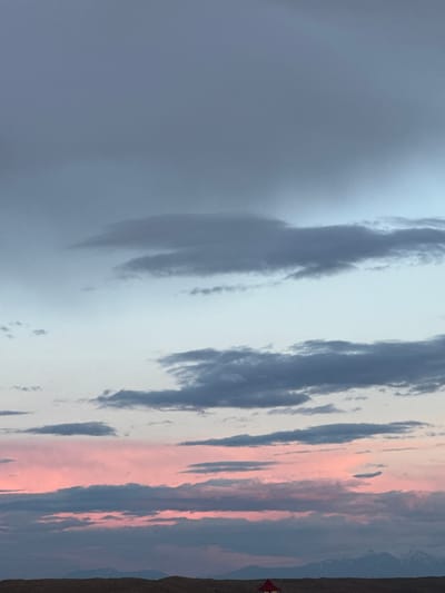 The image shows a landscape at twilight or sunrise, likely in a rural area. Predominantly, the sky is filled with clouds of varying shades of gray, white, and pink, suggesting the presence of a colorful sunset or sunrise. On the right side of the image, a small patch of land is visible, with some scrub vegetation. In the distance, there's a small red object that could be a building or a vehicle. The overall setting seems to be an open space with a dramatic sky. There is no way to know if this scene took place in Anipemza, Armenia without further context.
