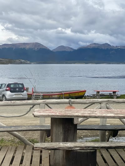 The image depicts a static, outdoor scene in Puerto Almanza, Argentina, presenting a view across a body of water toward distant mountains under an overcast sky.
In the foreground, a rustic wooden fence and railing system spans the width of the frame. A weathered wooden picnic table with a faded red tabletop is positioned in the immediate foreground, partially obscured by the fence. A thick, dark rope is wrapped around a fence post, secured to an orange, spherical buoy hanging behind the top rail. The ground in the foreground is composed of dirt and gravel, with wooden planks visible at the very bottom.
The middle ground features a shoreline with a silver compact hatchback car, identified as a Fiat with license plate "AD 094 PB," parked on the dirt. To its right, a weathered wooden boat, painted yellow on the hull and red on the gunwales, rests on the shore. The numbers "0709-H" are visible in white on the boat's side. Wooden steps lead up to the boat. A cluster of bare, pale branches stands vertically near the boat. Further to the right along the shoreline, two additional rustic wooden picnic tables with benches, featuring faded red tops, are situated on a patch of sparse green grass.
The background consists of a wide expanse of calm, grey-blue water. Beyond the water, a range of large mountains with varying peak formations rises, their lower slopes covered in dark, dense vegetation, contrasting with lighter, rockier upper sections. The sky above is entirely overcast with a dense layer of grey and white clouds. No people or active interactions are observed in the scene.
