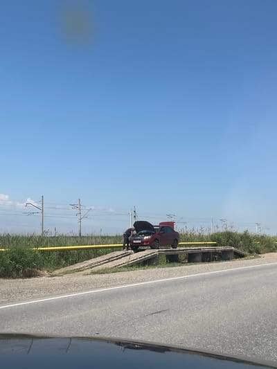 In Surenavan, Armenia, a dark red car is parked on the side of a road, with its hood open. Two individuals are visible near the car, appearing to be attending to it. The scene is set on a bright, clear day, suggested by the vivid blue sky. The car is situated on a paved road, bordered by a grassy verge and a concrete barrier. Beyond the verge, tall reeds or grass are growing, and power lines with poles are visible in the distance, indicating a semi-rural or roadside setting. The lighting suggests it's daytime. A portion of another dark-colored vehicle is reflected in the foreground, possibly the one from which the photo was taken. The license plate of the red car is partially visible and reads "M 609 YX 64". The overall mood appears to be one of a vehicle breakdown or maintenance.