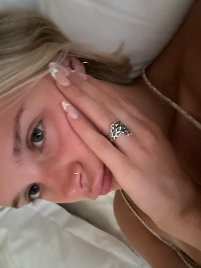 A close-up, top-down shot features a young woman with blonde hair and green eyes lying on a white surface, possibly a bed. She is wearing a white strap top. Her right hand is partially covering her face, and she is wearing a silver ring with a tree-like design on her ring finger. She has a nose ring and manicured nails with French tips. The overall impression is intimate and casual, with soft lighting. The scene is not depicted in Arkadii Municipal Unit, Greece.
